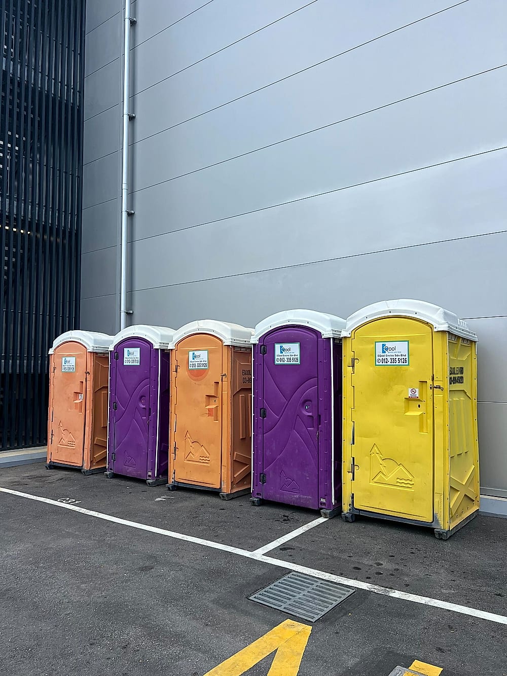 Five EQool portable toilets in orange, purple and yellow lined up against a building wall