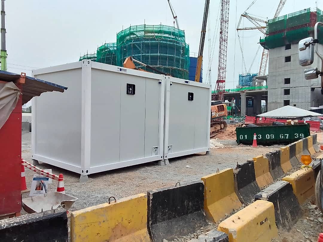 Two white container toilet cabins installed on gravel beside cranes and a building under construction