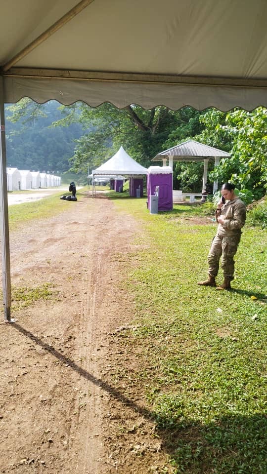 Soldier standing on the grass path at the US Army training camp, with purple EQool portable toilets and white tents behind her