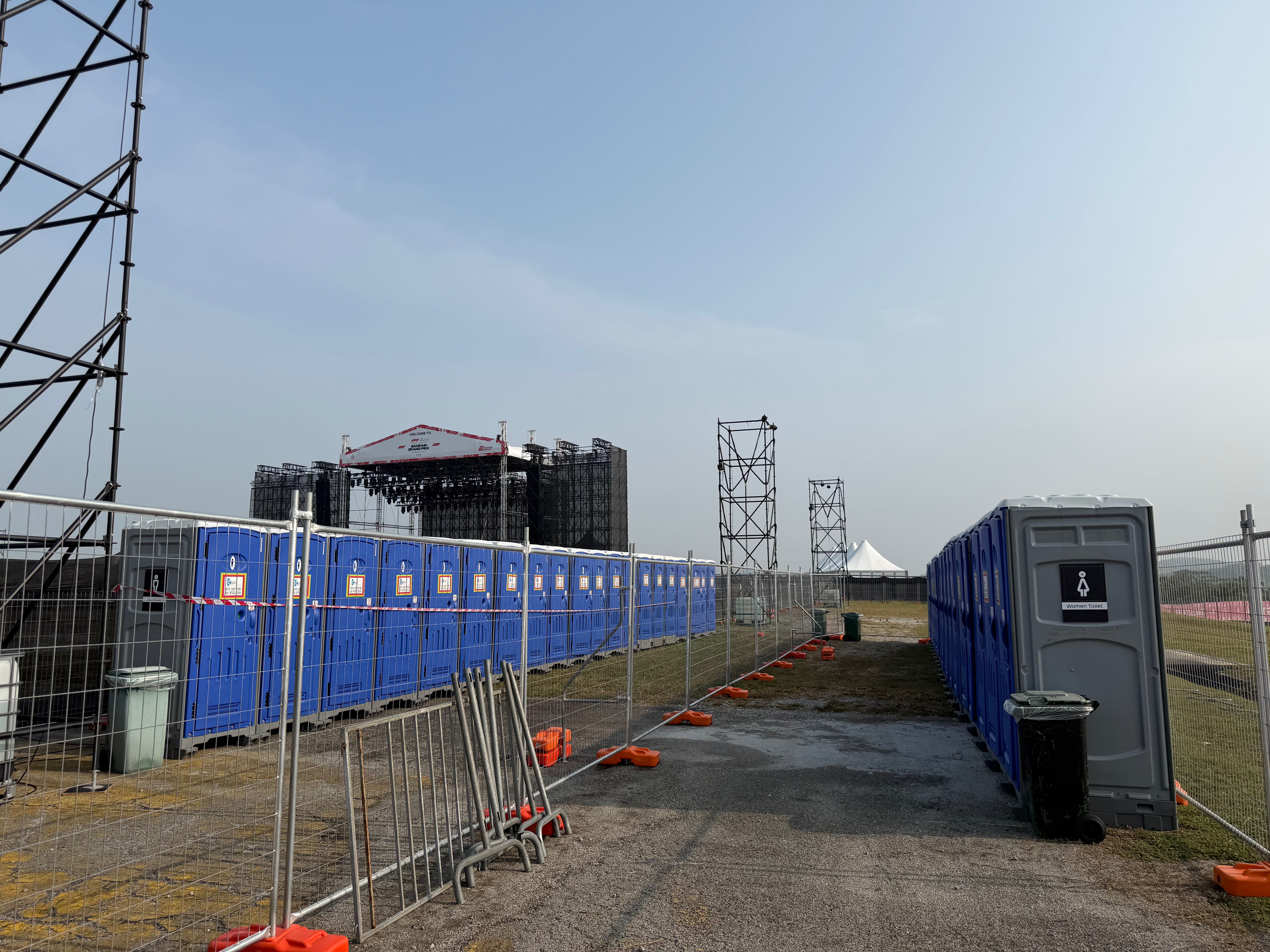Fenced rows of blue and grey EQool portable toilets with the Bahrain Grand Prix stage in the background