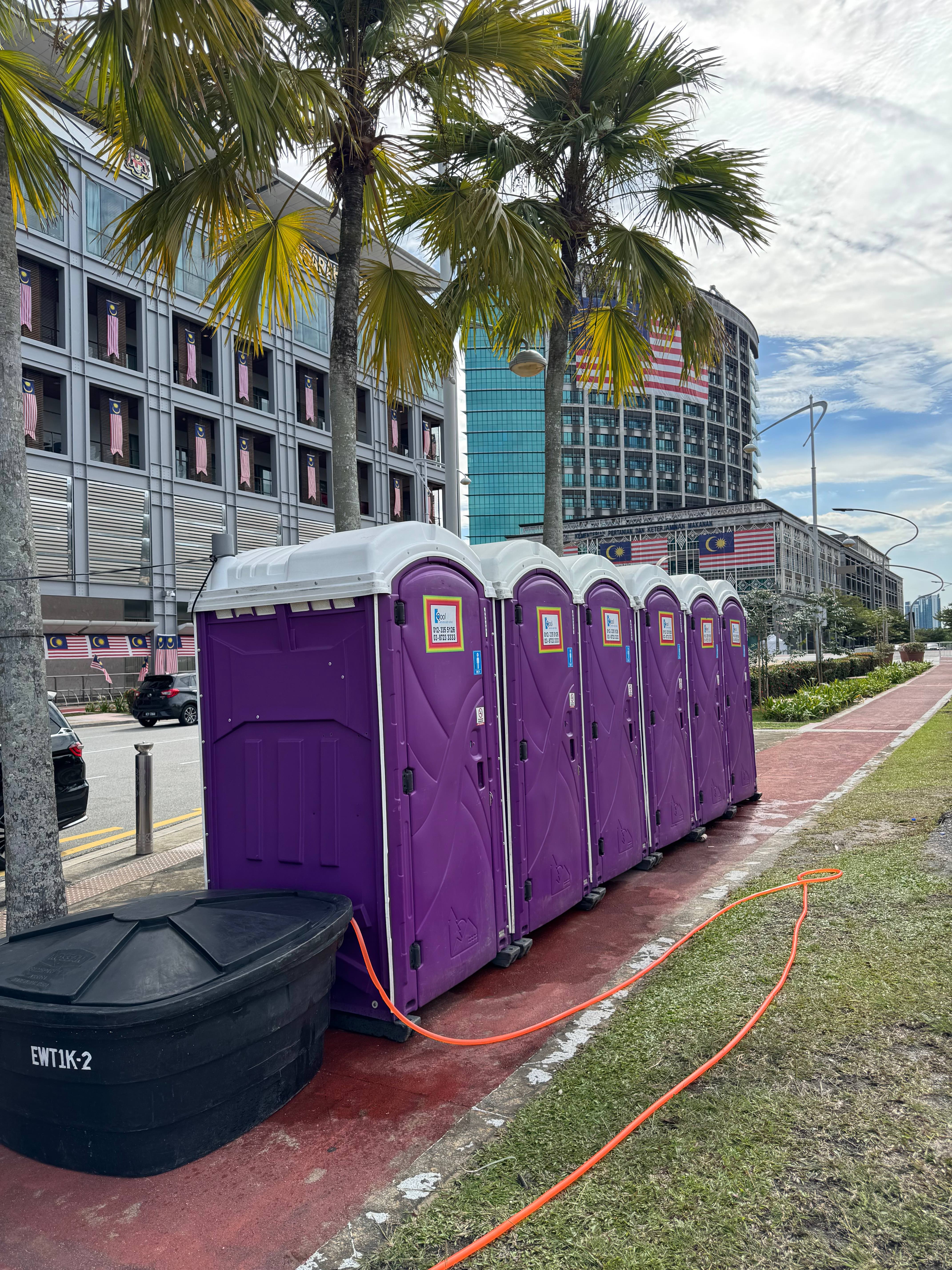 Purple EQool portable toilets connected by an orange hose to a black water tank, with Malaysian flags on the buildings behind