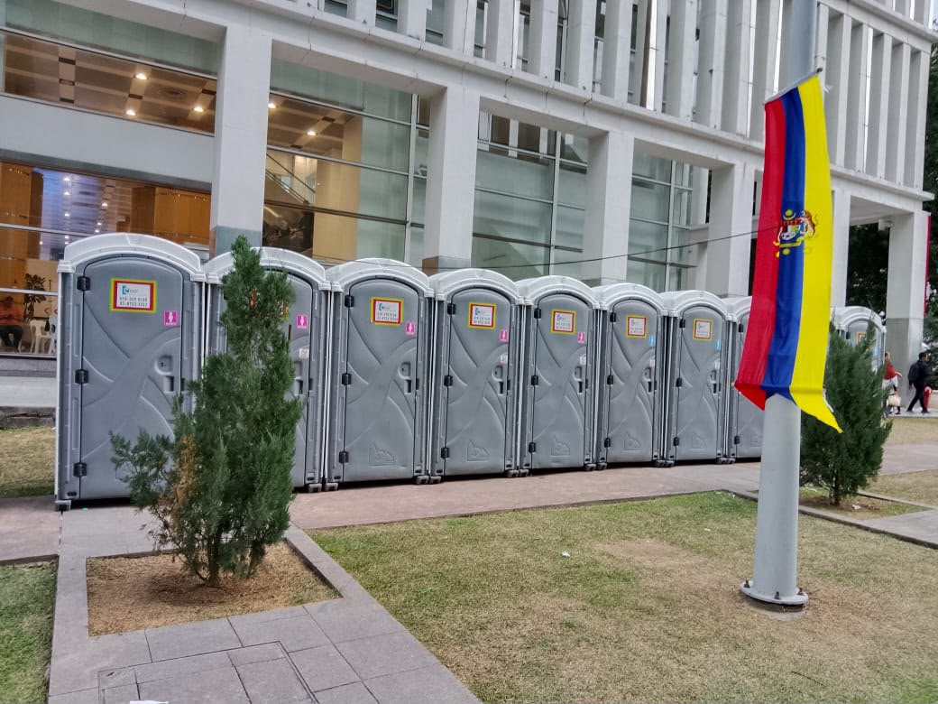 Row of grey EQool portable toilets on a paved path beside a city building, with small trees and a flag banner in the foreground