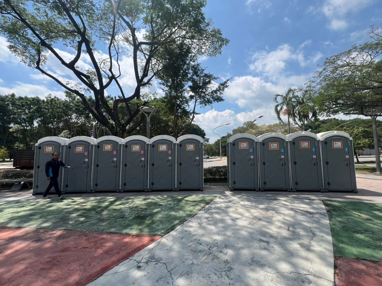 Grey EQool portable toilets in two groups, ladies and gents, in a tree-shaded park with a crew member walking past
