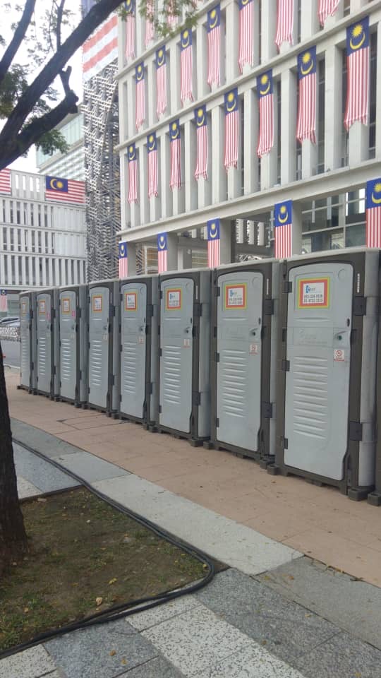 Grey EQool portable toilets lined up beside a building covered with Malaysian flag banners