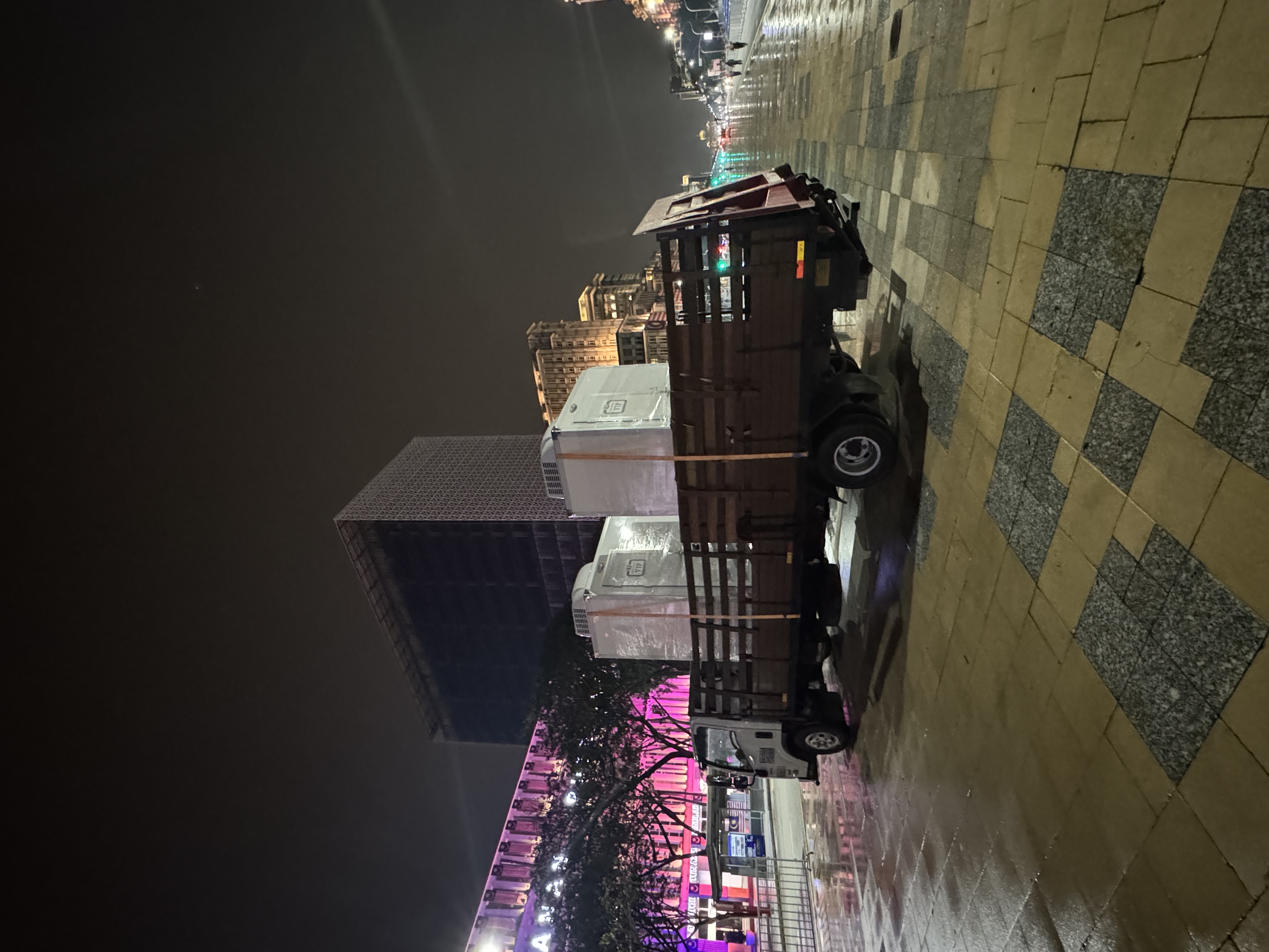 Lorry carrying two wrapped VIP cabin toilets through a wet plaza at night during delivery for Hari Kebangsaan 2025